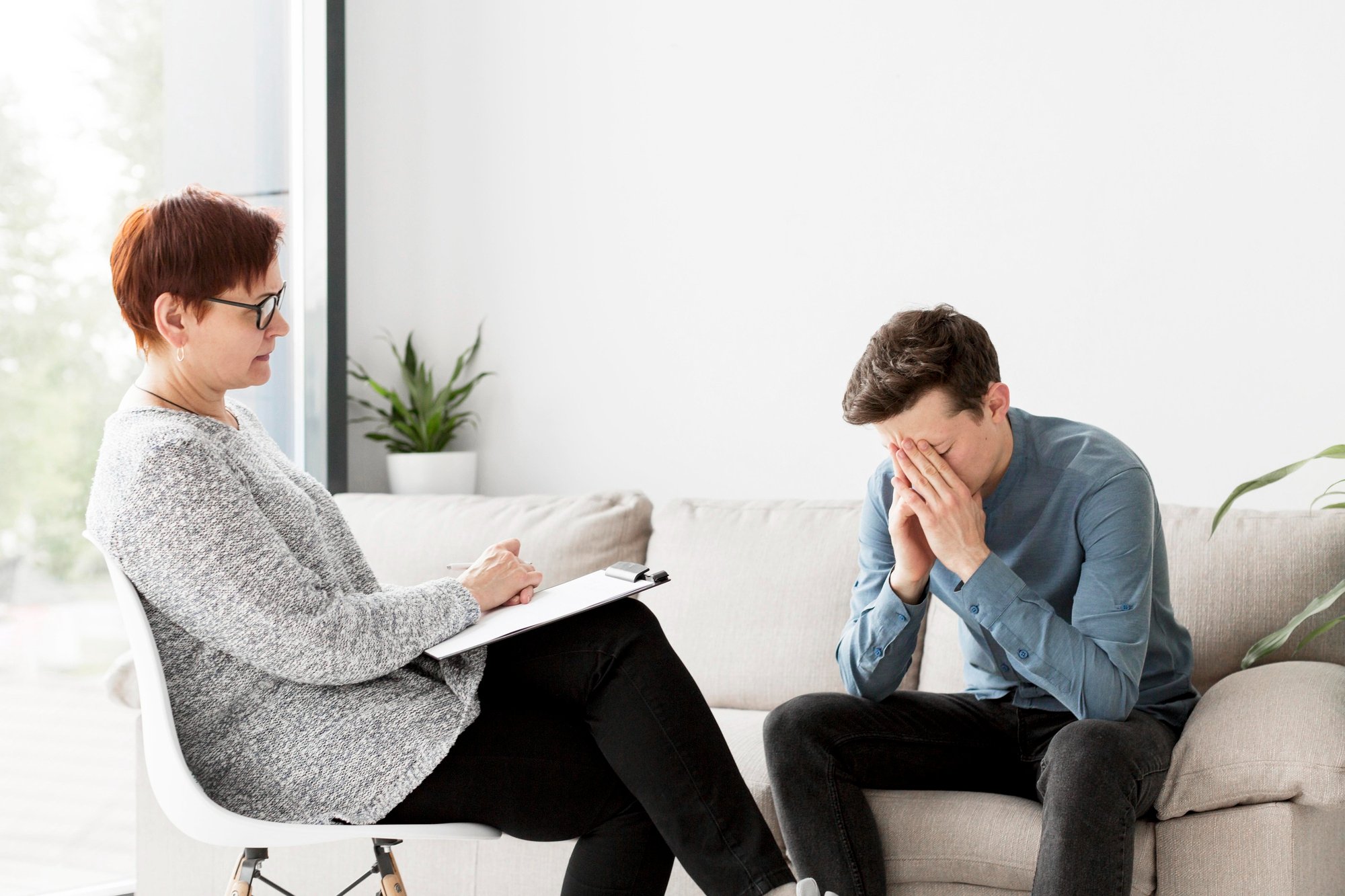 Counselor records observations while a client on a couch covers his face in distress.