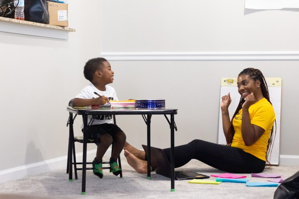 Child at a table doing schoolwork while a woman sits on the floor encouraging him.