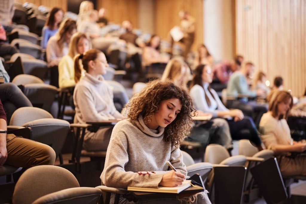Curly-haired student writes notes in a busy lecture hall.