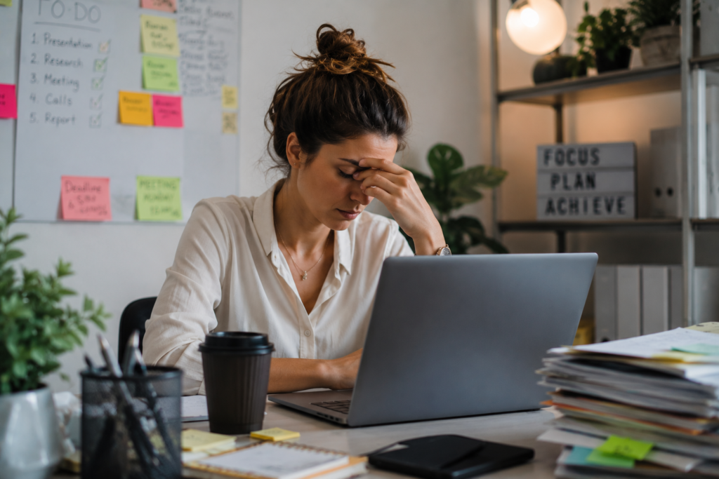 High-achieving adult woman showing signs of ADHD burnout while working late at a desk with a laptop, paperwork and sticky notes in a realistic office environment.